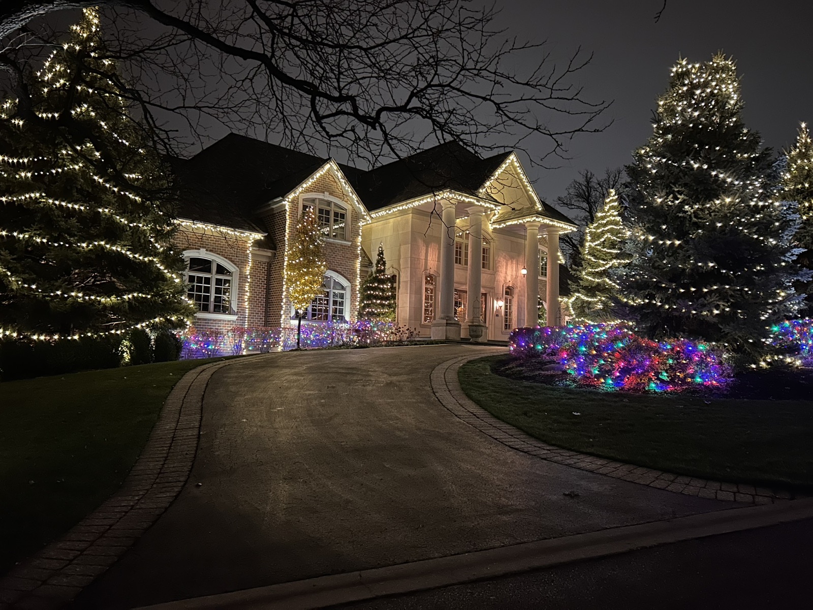 Estate home with warm white roofline lighting and two large wrapped evergreens