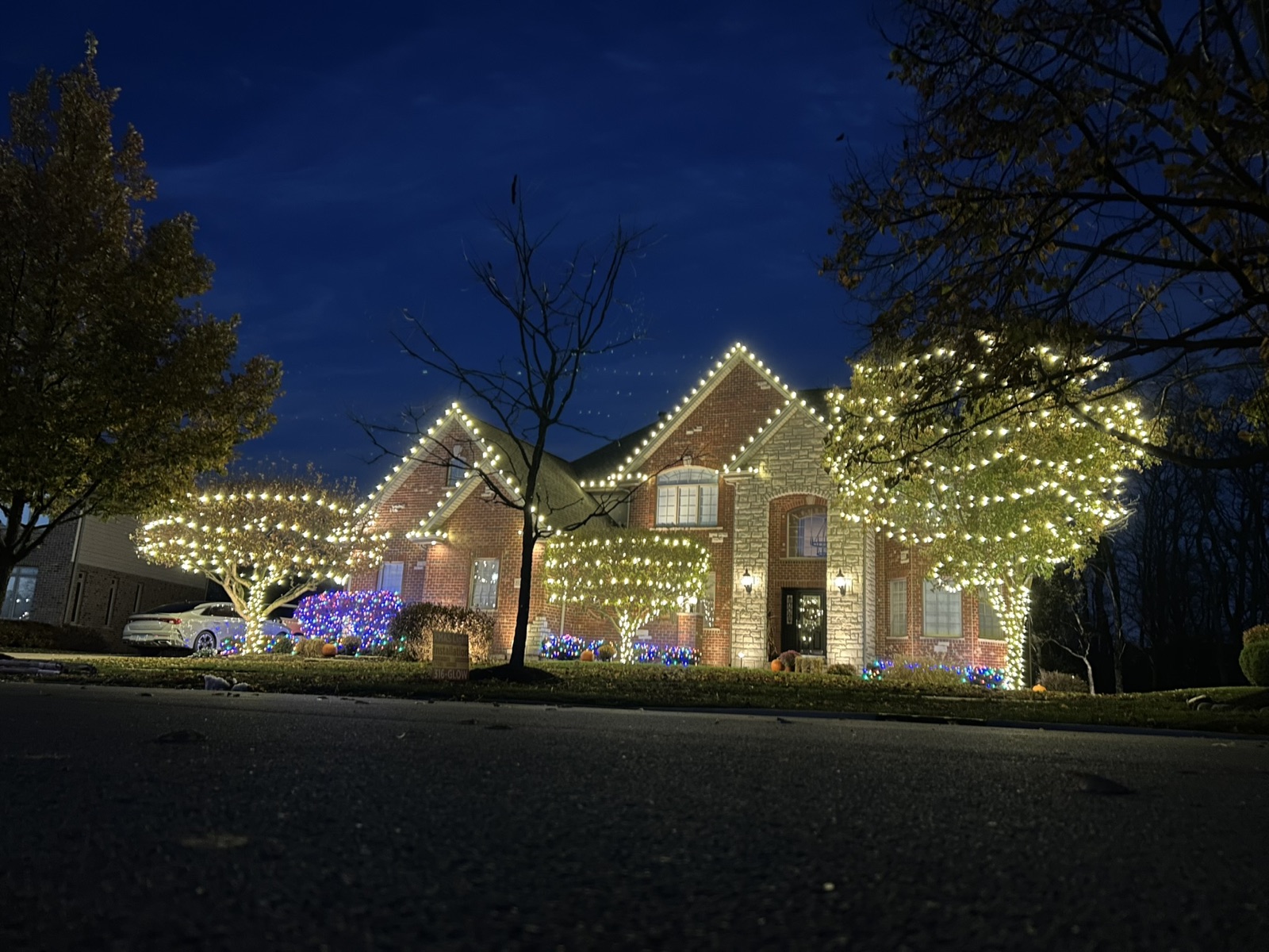 Large brick home lit with warm white roofline lights and wrapped trees at dusk